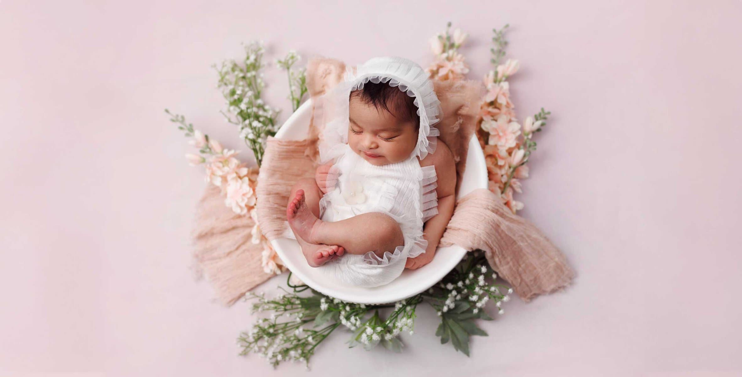 A peaceful newborn, dressed in white, rests in a bowl adorned with pink fabric and flowers, capturing the essence of newborn photography against a soft pink backdrop.