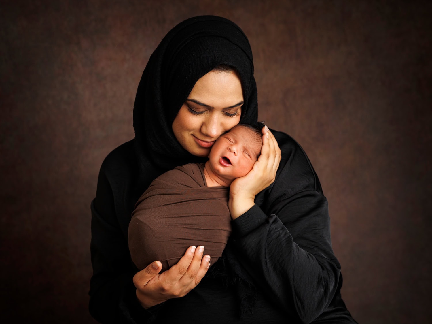 A woman in a black headscarf cradles her sleeping newborn, wrapped in brown fabric, against a dark backdrop. This serene moment captures the essence of newborn photography.