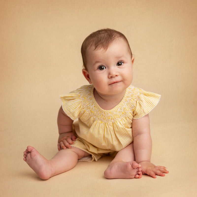 A sitter baby in a yellow outfit sits on a beige background, gazing forward with a neutral expression.