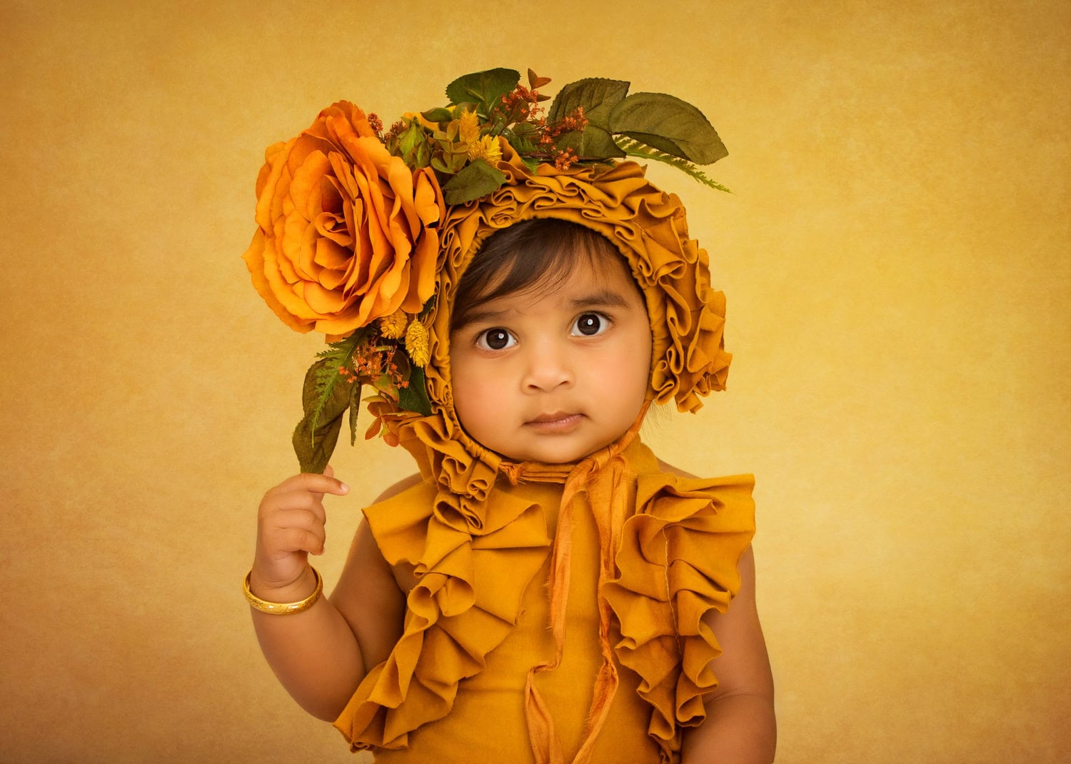In a charming sitter baby portrait, a young child is dressed in a yellow outfit with ruffles, complemented by a large headpiece adorned with orange flowers on a matching yellow background.