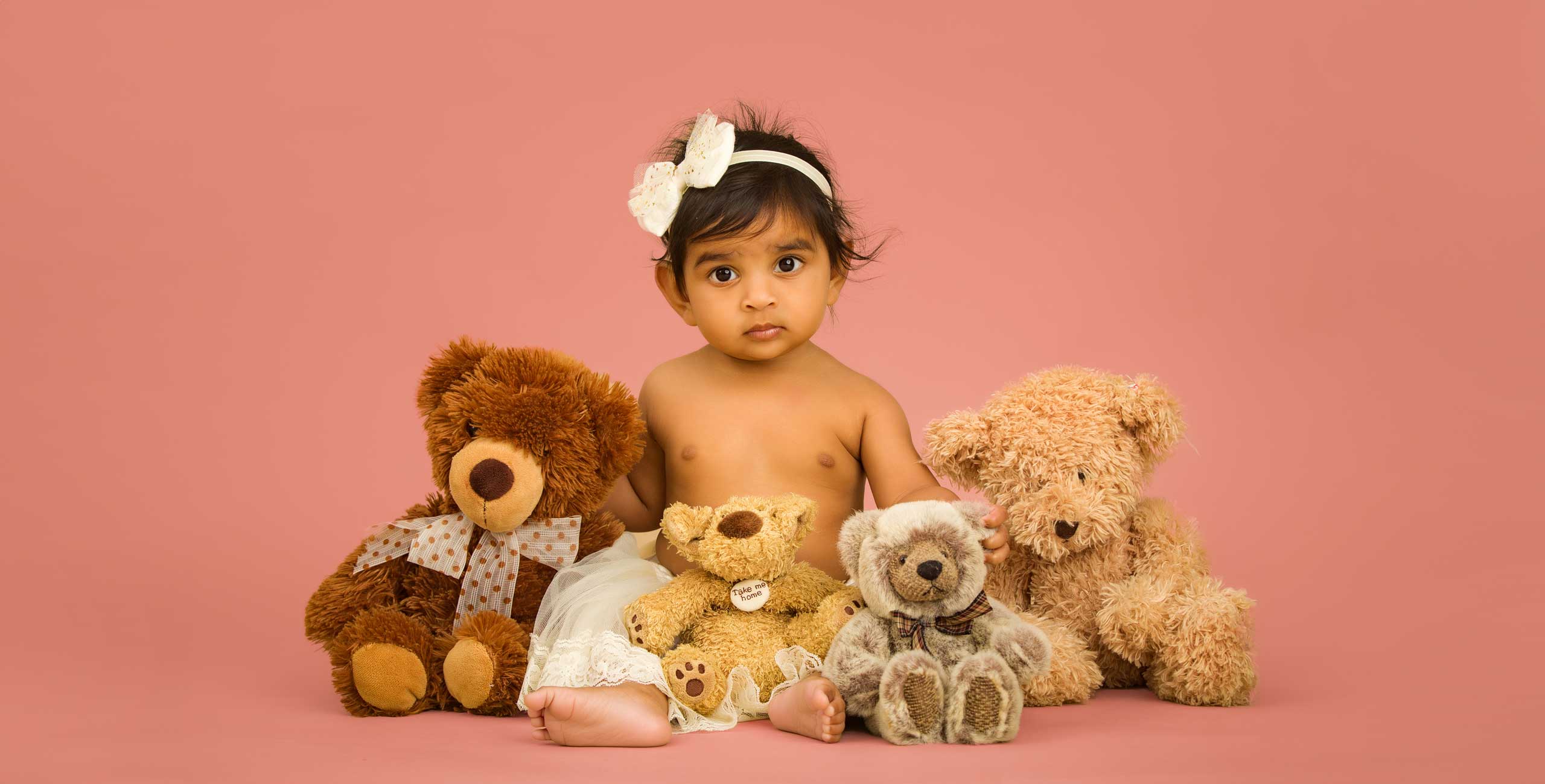 A sitter baby in a white headband sits on a pink background, surrounded by four plush teddy bears.