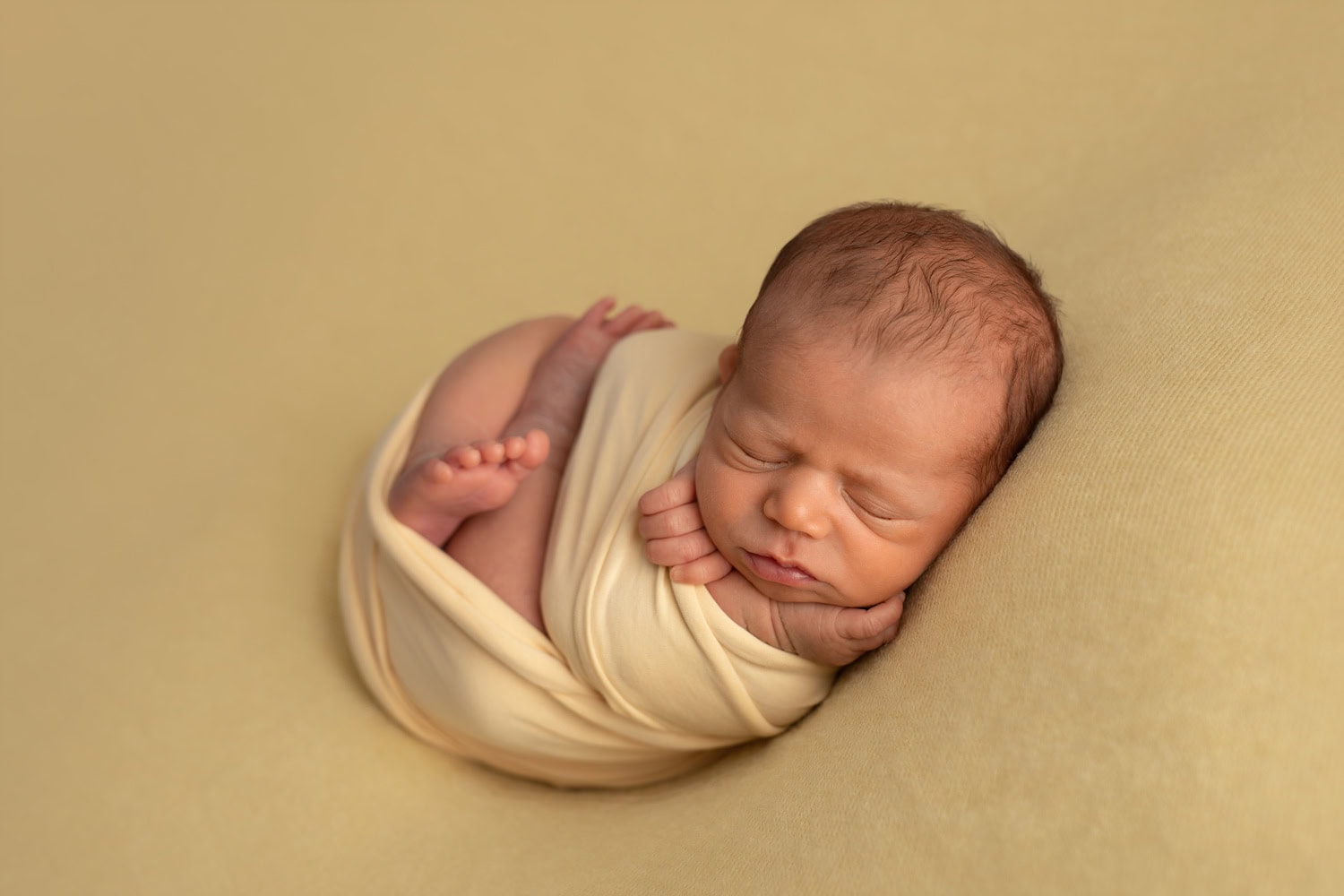Newborn photography captures a baby sleeping peacefully on a yellow blanket, wrapped in a soft, light yellow swaddle.