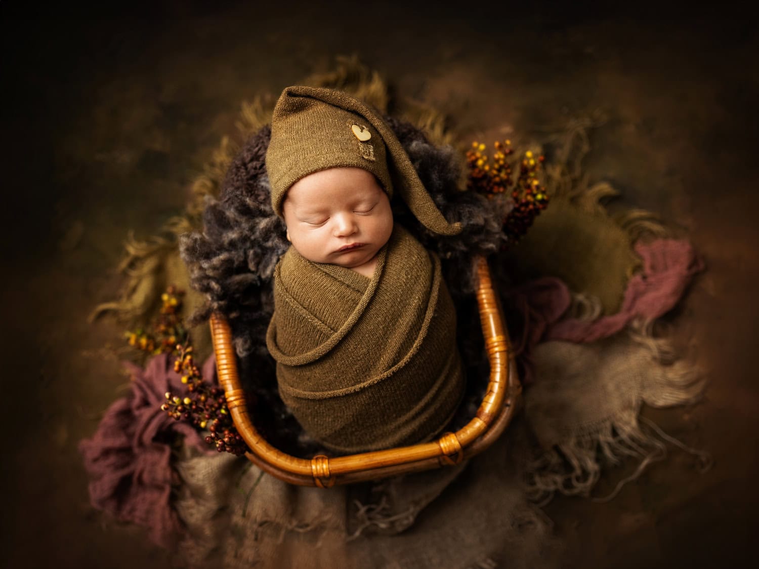 Newborn photography captures a sweet scene: a baby swaddled in olive green fabric, complete with a matching hat, sleeps peacefully in a basket. The surrounding textured cloth and small floral accents enhance the charming composition.
