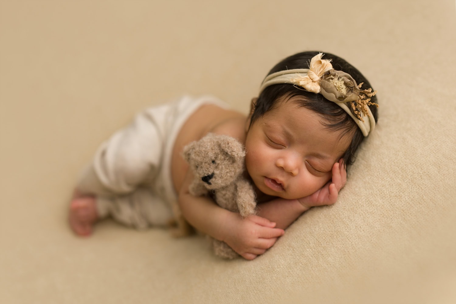 A newborn from 2025 sleeps peacefully on a beige surface, adorned with a delicate floral headband and cozy white pants, cradling a small teddy bear.