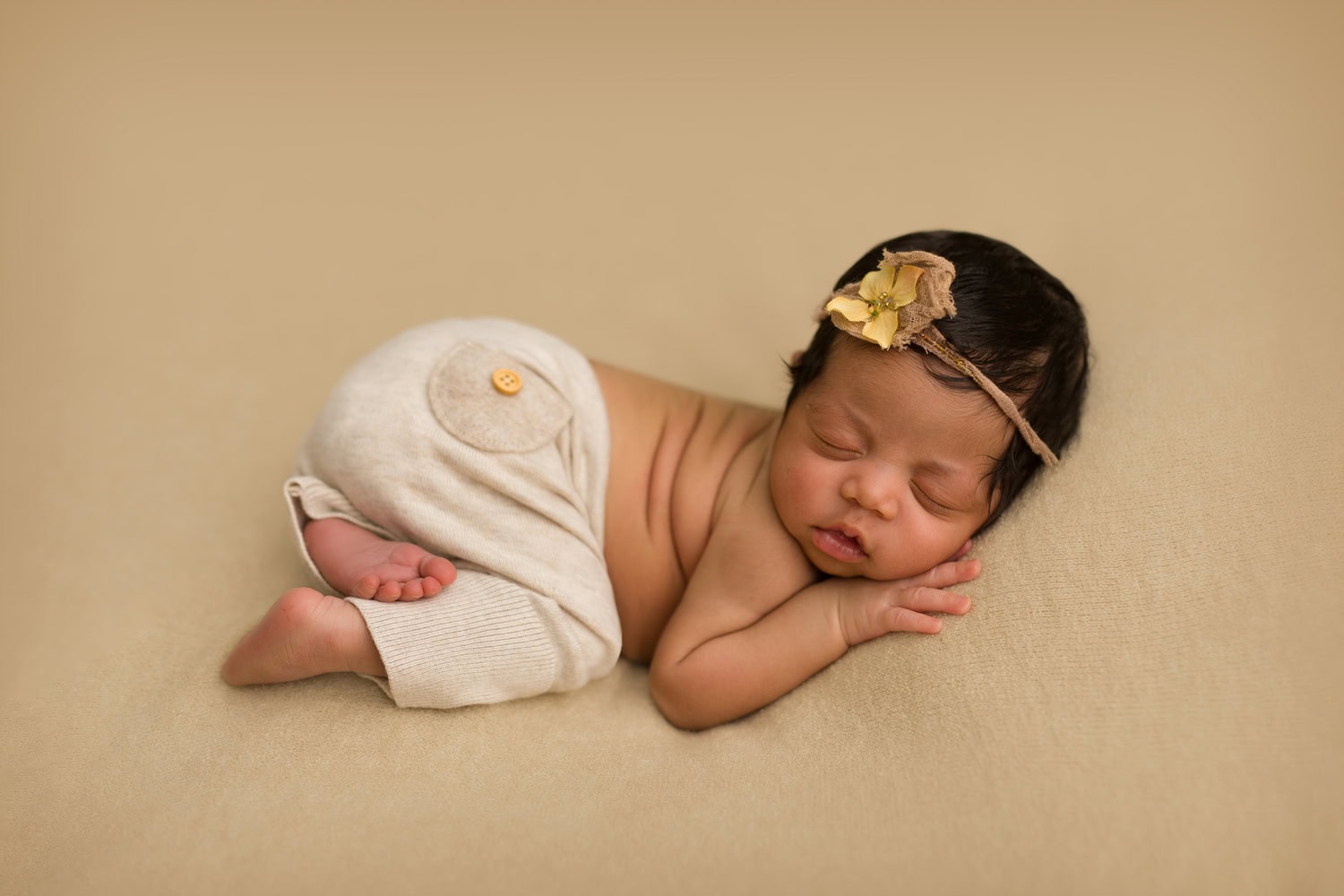 A peaceful newborn drifts to sleep, dressed in beige pants and a delicate headband adorned with a fabric flower, resting on a soft beige surface.