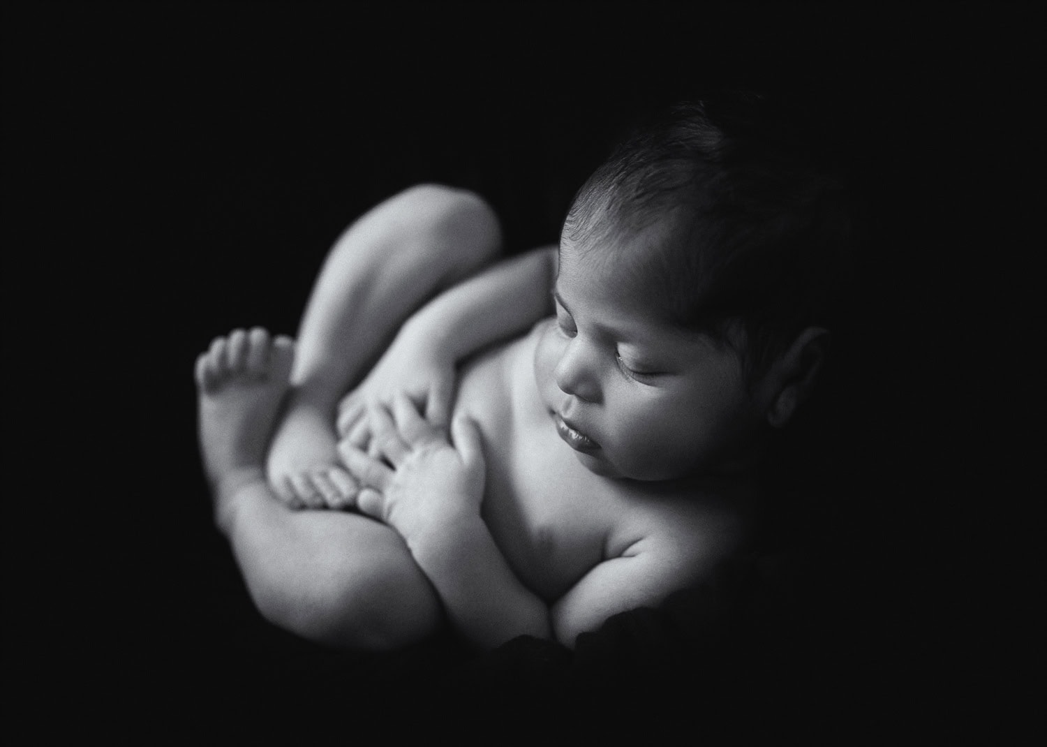 In a timeless black and white photo from 2025, a sleeping newborn baby rests curled up peacefully against the dark background.