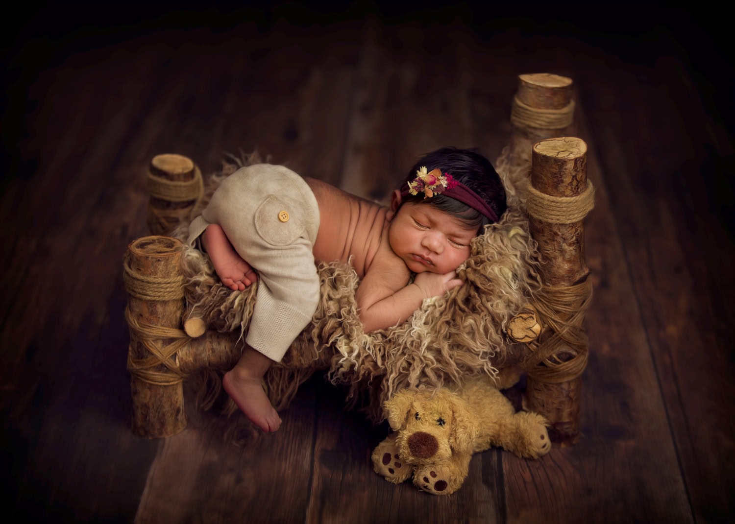 A newborn with a headband lies peacefully on a small wooden bed covered with a textured blanket, embodying serenity. Nearby, a plush toy dog rests by the babys feet, completing this timeless scene poised to inspire in 2025.